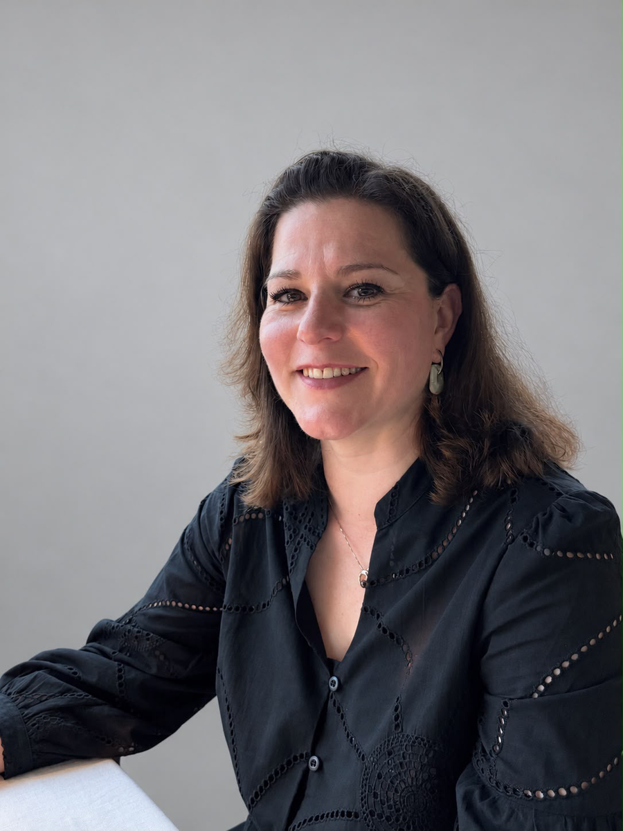 Kim Verlot. A smiling woman with shoulder-length brown hair wearing a black eyelet blouse, seated against a plain light gray background.